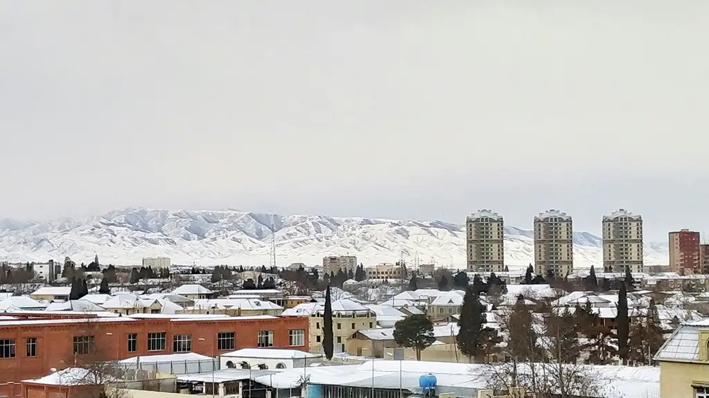 Gence'nin karla kaplı görünümü; alçak yapıların arasında yükselen birkaç kule blok ve arkada karlı dağ sırası.