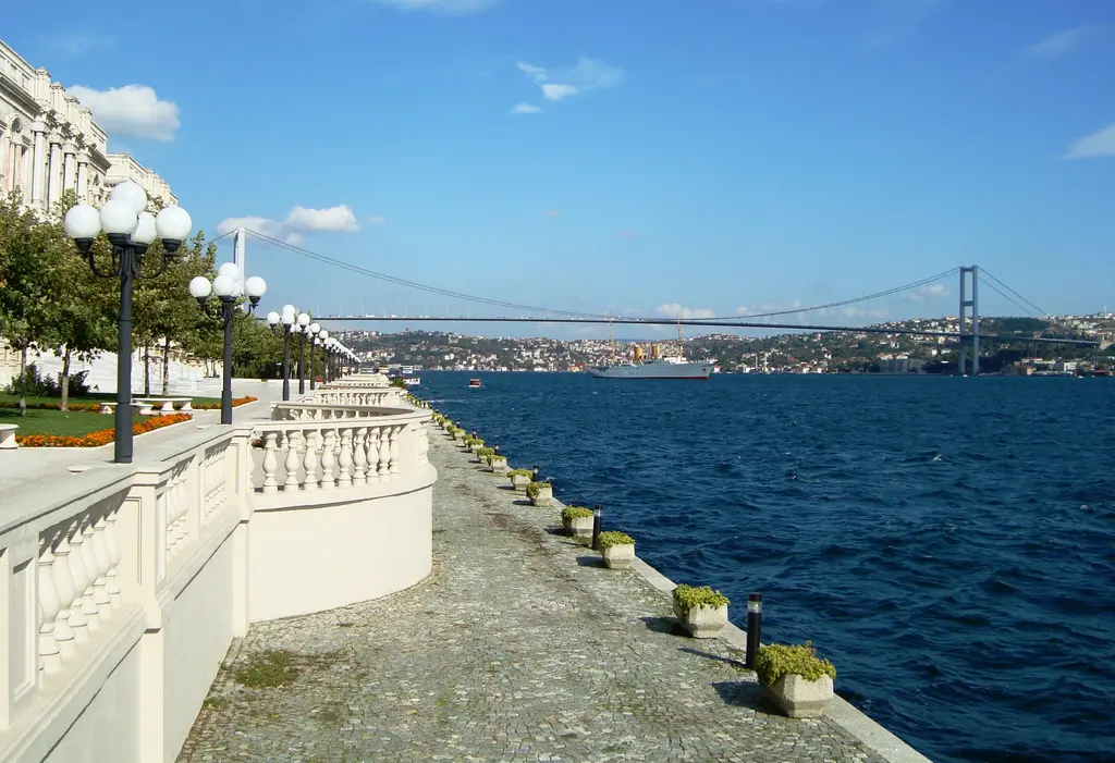 Çırağan Sarayı'nın rıhtımı; uzun mermer korkuluk, fenerler, sağda Boğaz'ın mavi suyu ve uzakta köprü.