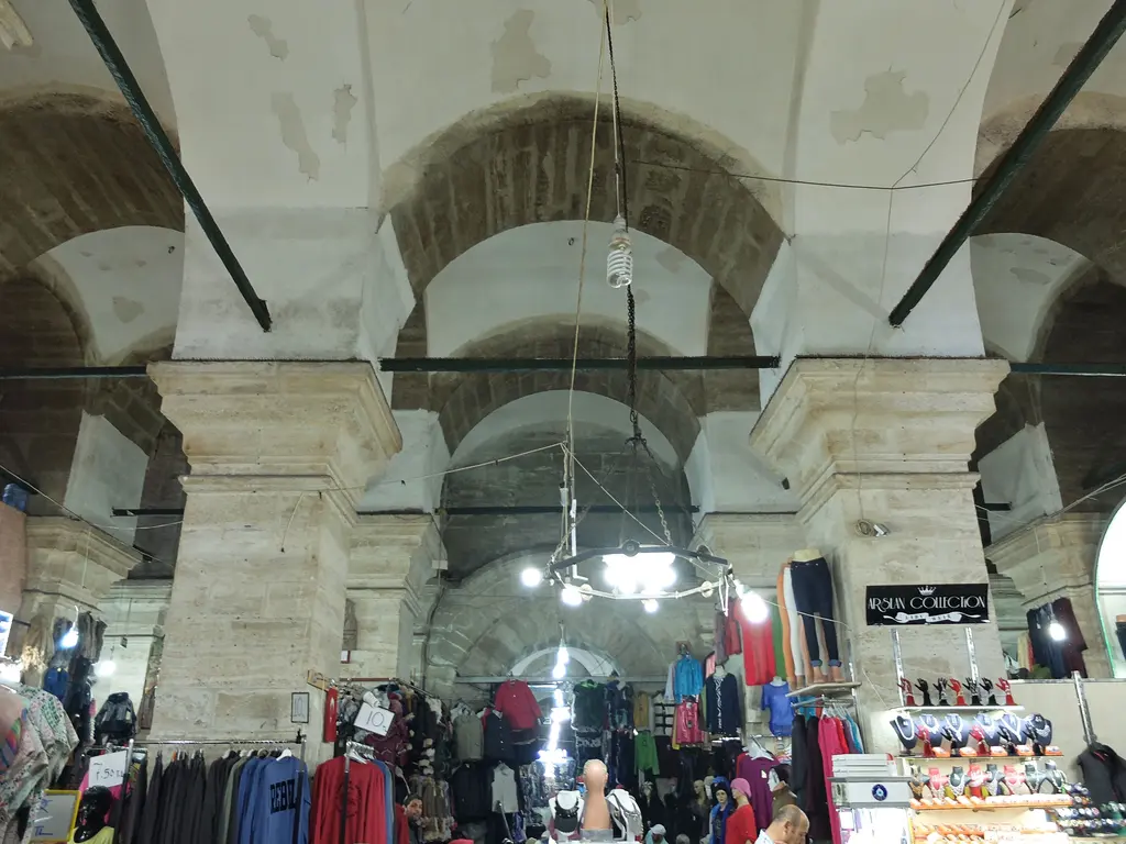 Laleli Camii'nin alt katı; kalın payelerin taşıdığı tonozlu mekân, aralara kurulmuş tekstil dükkânları ve askılarda giysiler.
