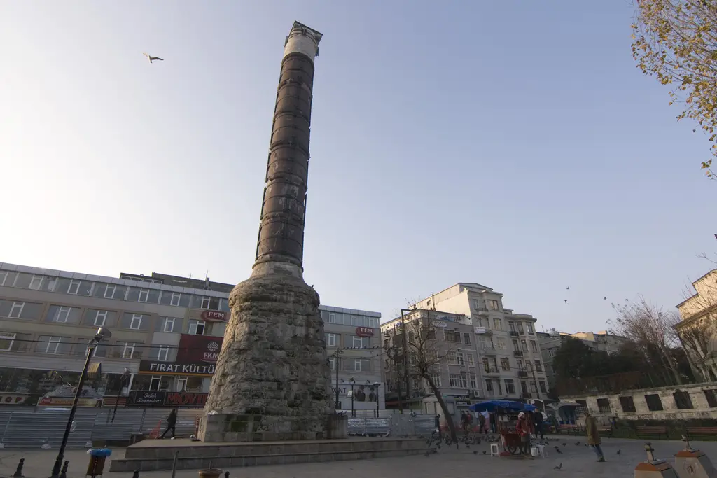 Çemberlitaş'ta meydanın geniş açısı; solda sütun, sağda çok katlı cadde binaları, düzenlenmiş taş zemin ve kapalı gökyüzü.