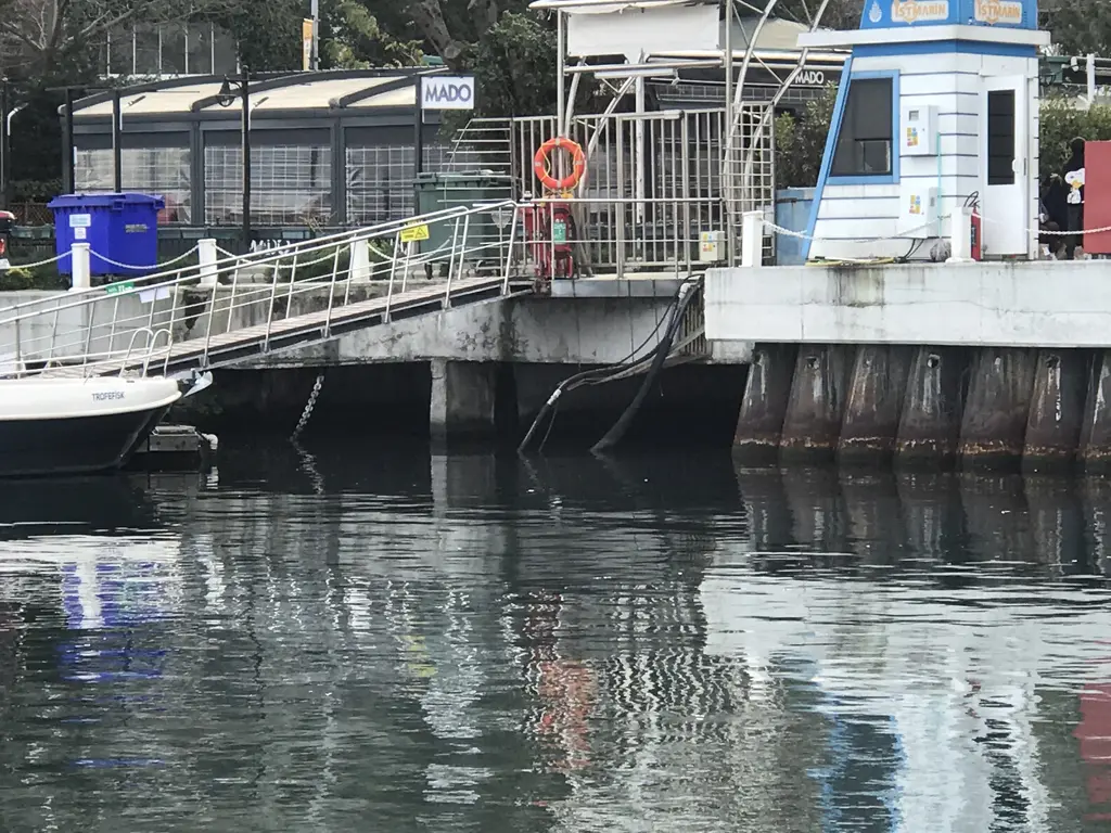 Tarabya İskelesi; suya uzanan rampa, korkuluklar, mavi beyaz kulübe, arkada kafe ve suda yansımalar.