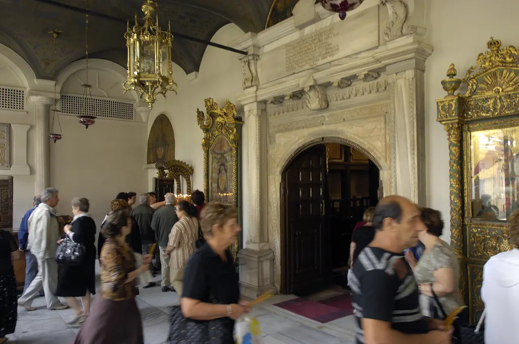 Aya Yorgi Kilisesi'nin narteksi; mermer söveli kemerli kapı, duvarlarda yaldızlı çerçeveli ikonalar, tonozlu tavan ve içeri giren ziyaretçiler.