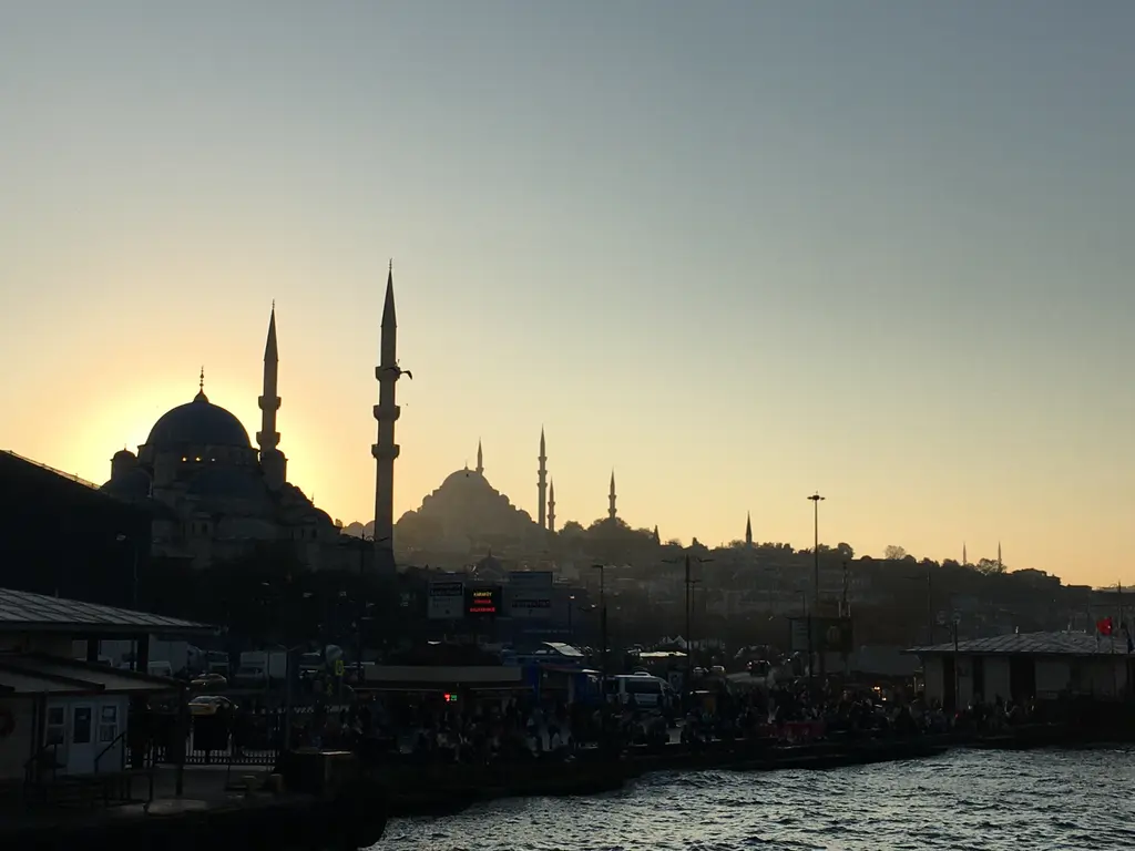 Gün batımında Yeni Cami ve Süleymaniye'nin siluetleri; turuncu gökyüzü, minareler, kıyıdaki yapılar ve su.