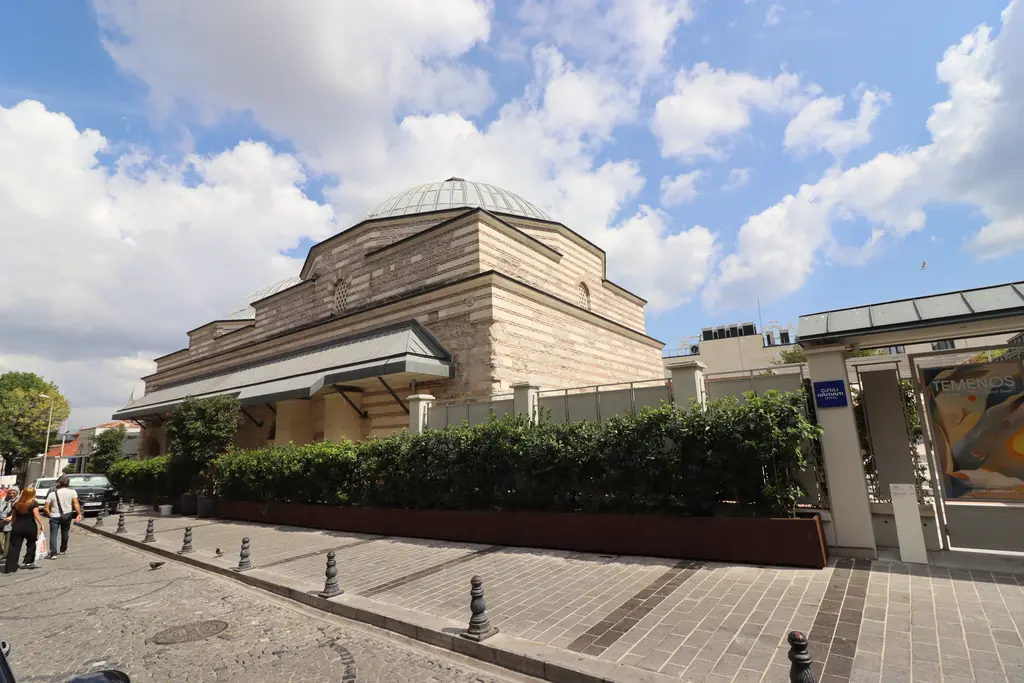 Zeyrek Çinili Hamam'a sokaktan bakış; almaşık taş ve tuğla örgülü kütle, kurşun kaplı kubbe, önde çit ve babalı kaldırım, yürüyen insanlar.