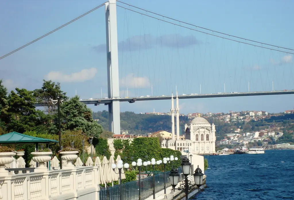 Çırağan Sarayı'nın kıyısından Ortaköy; sağda Ortaköy Camii, arkada asma köprü, önde ağaçlar ve mermer korkuluk.