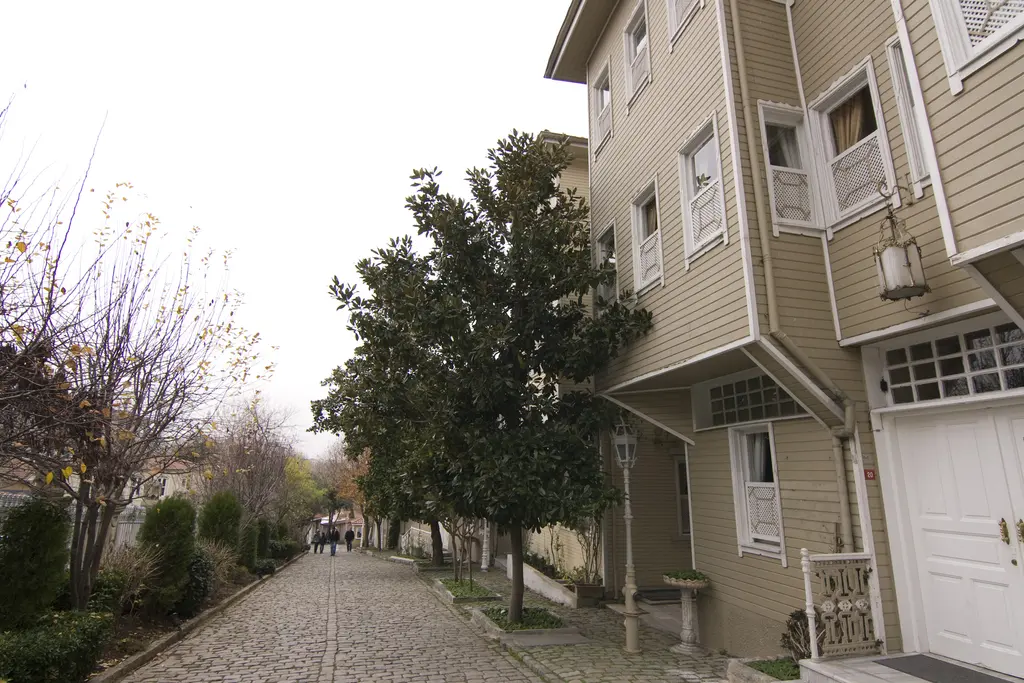 Kışın Soğukçeşme Sokağı; sağda beyaz garaj kapılı açık yeşil ahşap ev, önde yapraksız ağaç ve fener, taş döşeli yol boyunca yürüyenler.