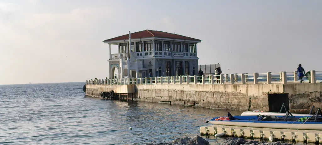 Moda İskelesi; denize uzanan taş yol, kiremit çatılı bina ve önde kıyıya bağlı küçük bir tekne.