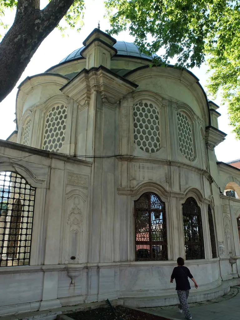 Şah Sultan Türbesi'nin hazireye bakan cephesi; dışbükey mermer duvar, petekli pencereler, kubbe ve yanından geçen bir kişi.