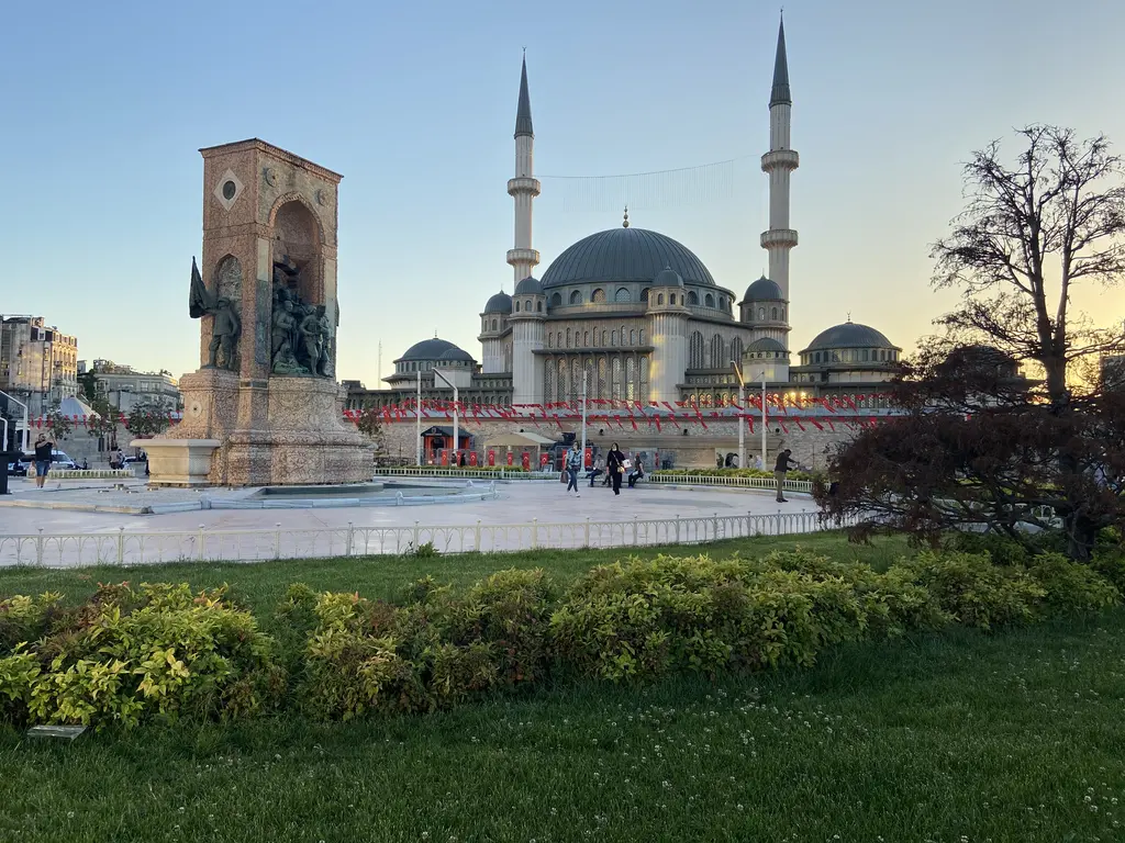 Gün batımında Cumhuriyet Anıtı ve arkasında Taksim Camii; önde yeşil çimler, çiçekli çalılar ve çıplak dallı bir ağaç.