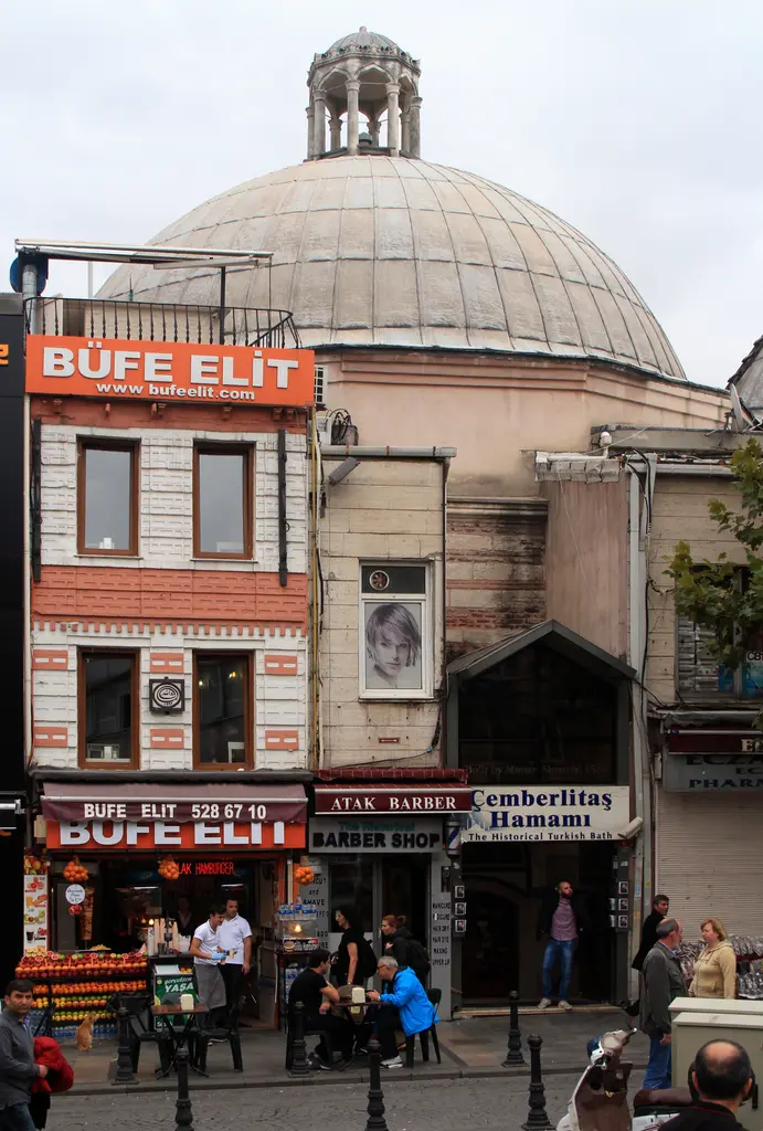 Çemberlitaş Hamamı'nın kubbesi dar bir açıdan; altında hamamın tabelası, büfe tabelalı köşe binası ve kaldırımda oturan insanlar.