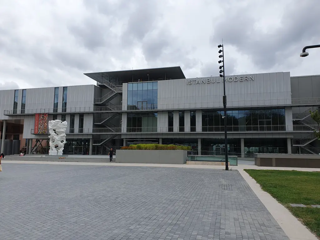 Bulutlu bir günde İstanbul Modern'in önden görünümü; geniş taş meydan, çim şerit ve binanın uzun cephesi.