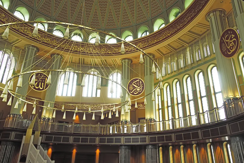 Taksim Camii'nin içi; kubbeyi taşıyan kemerler, pencerelerle dolu galeri katı, yuvarlak hat levhaları ve sarkan avize zincirleri.