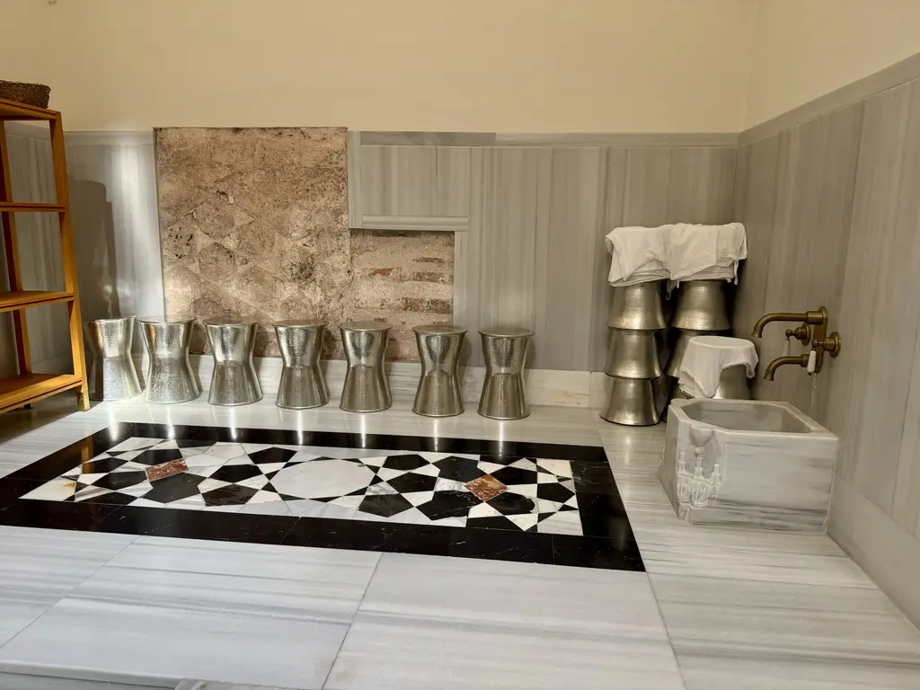 Çinili Hamam'da kurnalar; mermer tekneler ve pirinç musluklar, önlerinde sıra sıra metal hamam tası, yerde siyah beyaz geometrik döşeme.