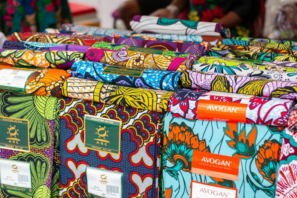 Batı Afrika pazarında üst üste yığılmış rengârenk mumlu baskı (wax print) kumaş topları; çiçek ve geometrik desenler, ABC ve Avogan marka etiketleriyle.