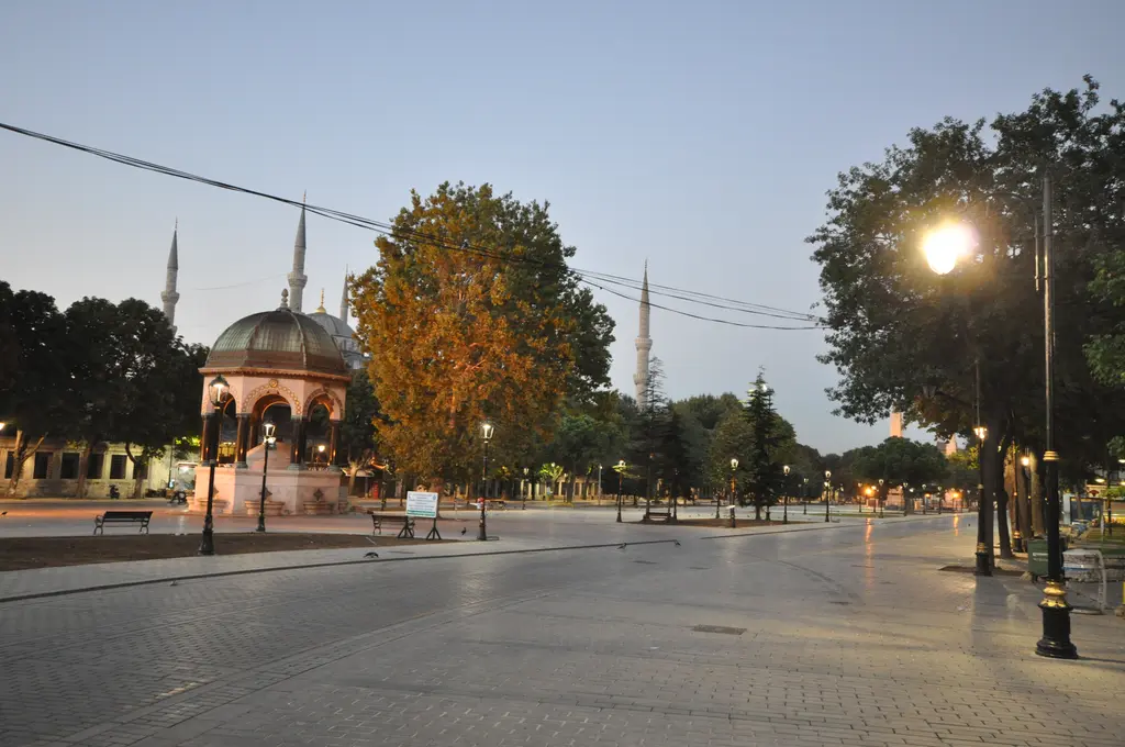 Sultanahmet'te akşamüstü hipodrom meydanı; solda kubbeli çeşme, ortada boş taş döşeme, sağda yanan sokak lambası ve arkada minareler.