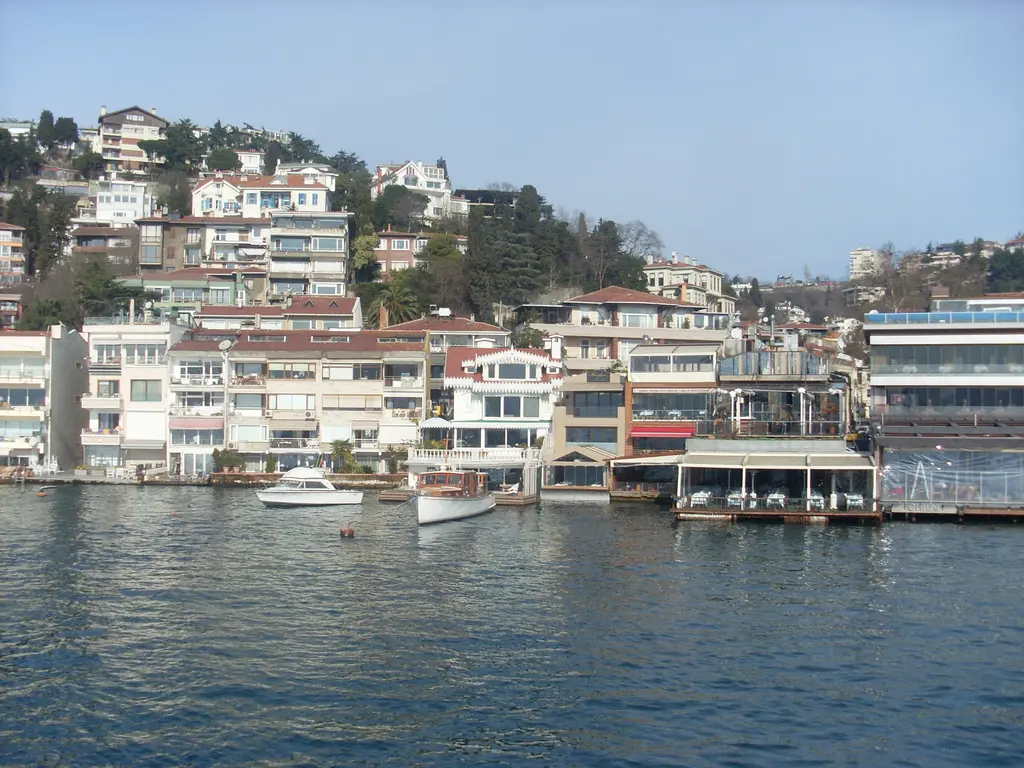 Bebek'te Boğaz'dan kıyı; suya inen beyaz ve renkli binalar, rıhtım lokantaları, tekneler ve yamaçta evler.