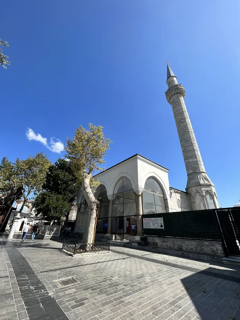 Fatih'te Koca Mustafa Paşa Camii'nin ön cephesi; camekânla kapatılmış üç kemerli son cemaat revağı, beyaz taş gövde, sağda tek şerefeli minare ve önde gri parke meydan.