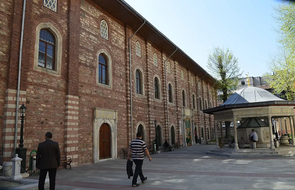 Arap Camii'nin avlusu; ağaçların arasında tuğla cephe, pencere sıraları, sağda şadırvan ve avluda oturan insanlar.