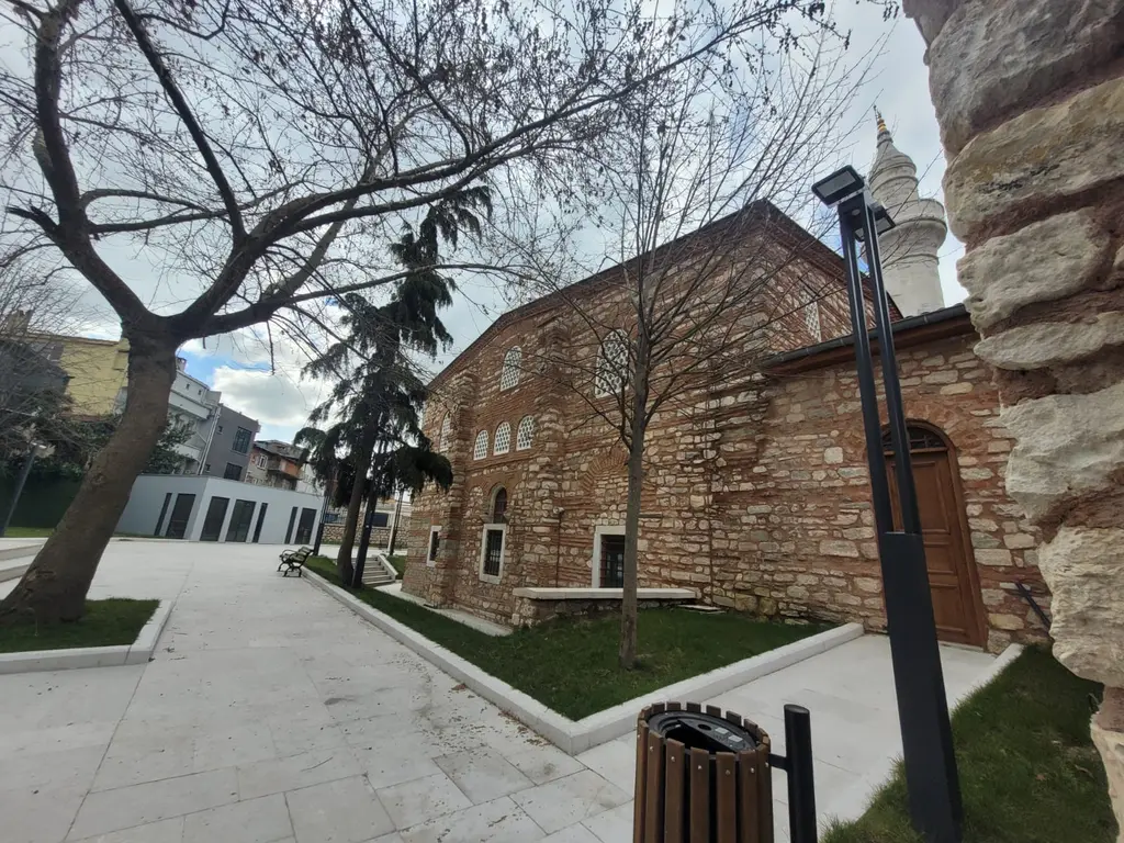 Atik Mustafa Paşa Camii'nin yanındaki yeni yaya alanı; beyaz taş döşeli yol, çimenlikler, çıplak dallı ağaçlar, sokak lambası ve arkada tuğla cami gövdesi ile minare.