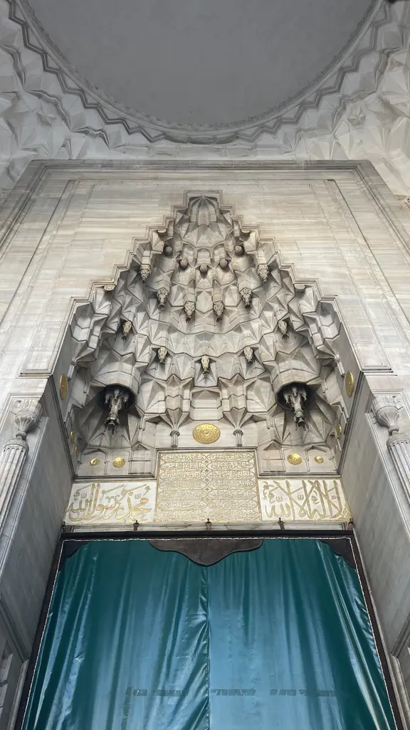 Şehzade Camii'nin taçkapısı tam alttan; beyaz mermerde derin mukarnas kavsara, yaldızlı göbekler, iki yanda kitabe panoları ve kapıyı örten turkuaz perde.