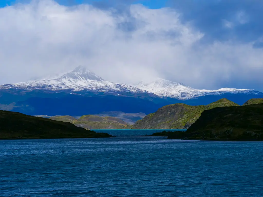 Patagonya'da bulutların arasından yükselen karlı bir zirve; önünde koyu tepelerle çevrili turkuaz-mavi buzul gölü, rüzgârlı ve bulutlu hava.