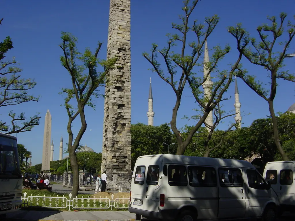 Örme Sütun'un taş gövdesi; önde budanmış ağaçlar ve beyaz minibüsler, arkada Dikilitaş ile Sultanahmet Camii'nin minareleri.