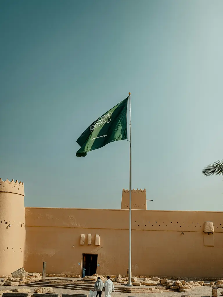 Riyad'da Masmak Kalesi; kerpiç sıvalı kalın duvarı, köşedeki silindirik burcu, koyu renkli ahşap giriş kapısı ve önünde dalgalanan Suudi Arabistan bayrağı.