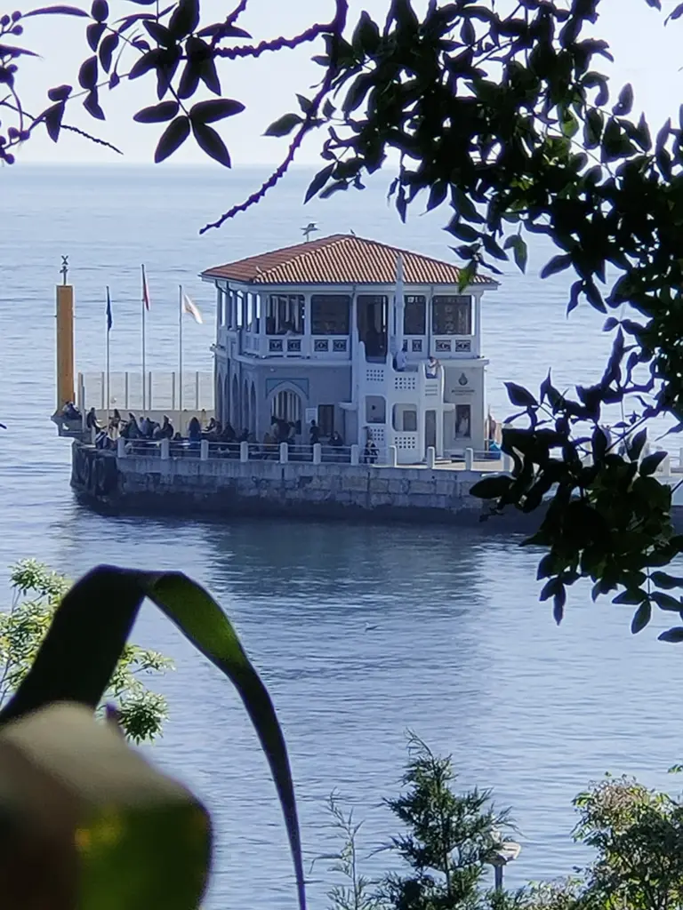 Moda İskelesi; ağaç dalları ve yaprakların arasından görünen bina, taş yol ve mavi deniz.