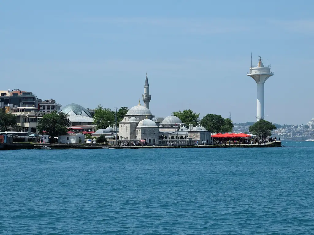 Şemsi Paşa Camii; kıyıda kubbeler ve minare, sağda beyaz bir kule, kırmızı tenteler ve mavi deniz.