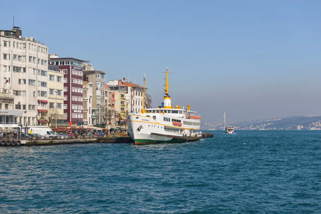 Karaköy iskelesine yanaşmış beyaz bir şehir hatları vapuru; kıyı boyunca renkli binalar, mavi su ve uzakta Boğaz.