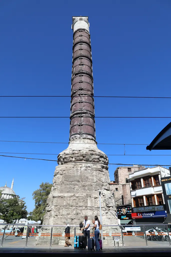 Çemberlitaş'ta sütunun dibi; kaba örgülü taş kaide, çevresindeki cam korkuluk, önünde duran ziyaretçiler ve bulutsuz mavi gökyüzü.