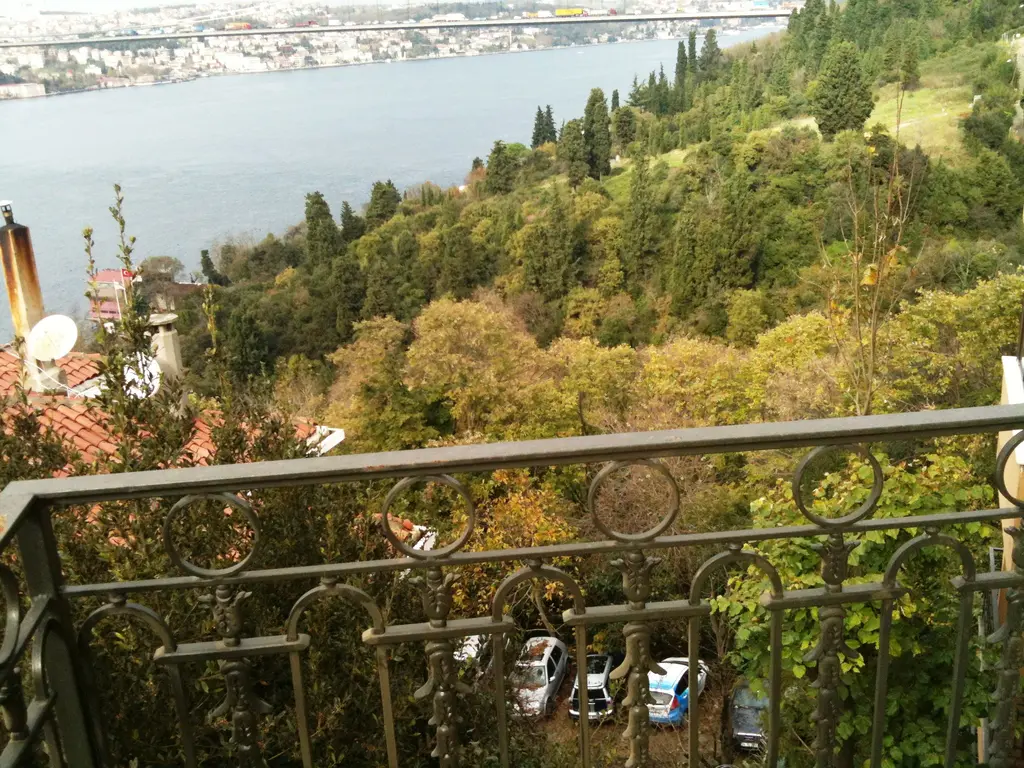 Mihrabat yamacında demir korkuluklu bir balkondan Boğaz'a inen ağaçlık sırt, arkada Boğaz'ın mavi suyu ve karşı kıyıdaki yerleşim.