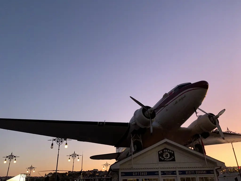 Rahmi Mustafa Koç Müzesi'nde Douglas DC-3; gün batımında pembe gökyüzüne karşı yükseltilmiş iki motorlu yolcu uçağı ve altında bir bina.