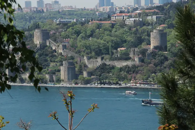 Sarıyer'de Rumeli Hisarı karşı kıyıdan; yeşil yamaca tırmanan kuleler ve surlar, mavi Boğaz ve önde çam dalları.