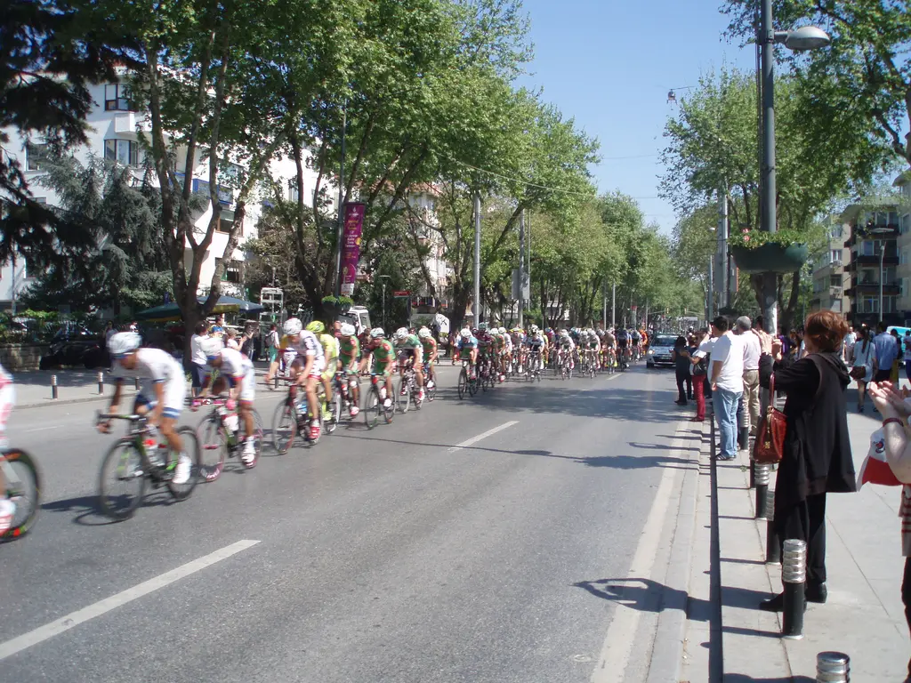 Bağdat Caddesi'nde bisiklet yarışı; caddeden geçen bisikletçiler grubu, kaldırımda izleyenler ve iki yanda çınarlar.