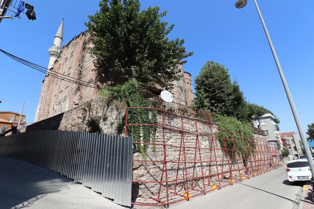 Gül Camii'ne dışarıdan bakış; yüksek taş ve tuğla gövde, minare, önde iskeleli dayanak duvarı ve ağaçlar.