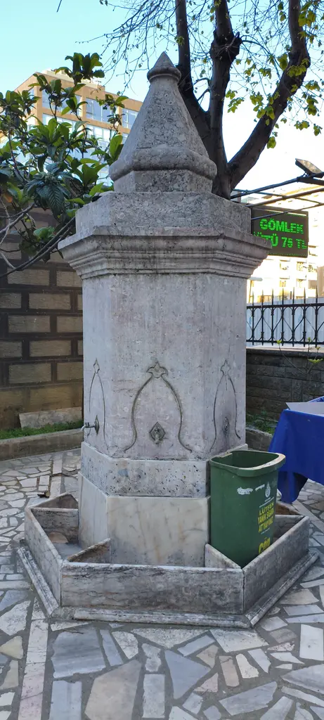 Göztepe'de Tütüncü Mehmet Efendi Camii'nin çeşmesi; sivri külahlı taş çeşme, taş döşeli avlu ve arkada ağaçlar.