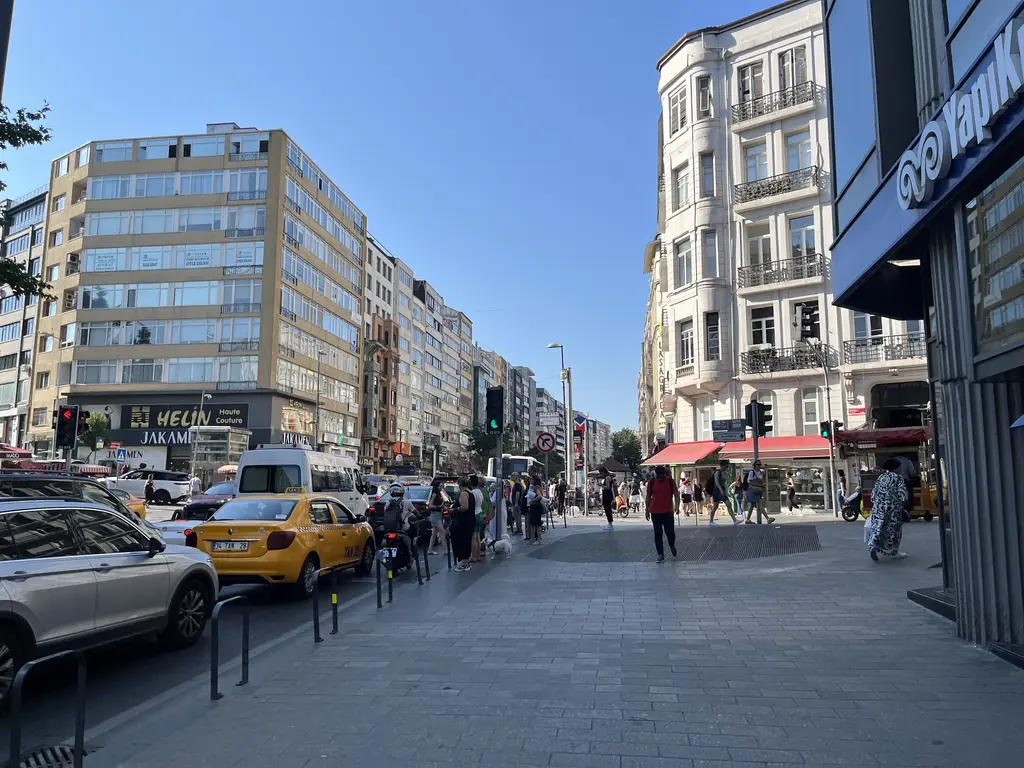 Osmanbey'de Halaskargazi Caddesi; geniş kaldırım, iki yanda eski ve yeni apartmanlar, dükkân tenteleri, sarı taksi ve yayalar.