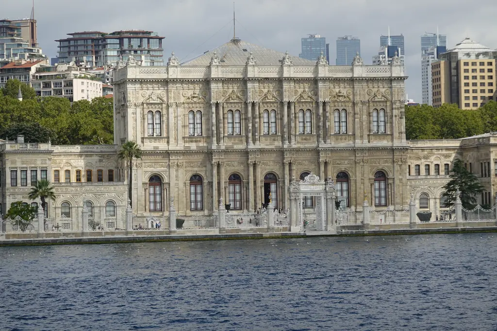 Denizden Dolmabahçe Sarayı; kıyı boyunca uzanan beyaz cephe, ortada süslü ana bölüm, arkada ağaçlar ve tepedeki gökdelenler.