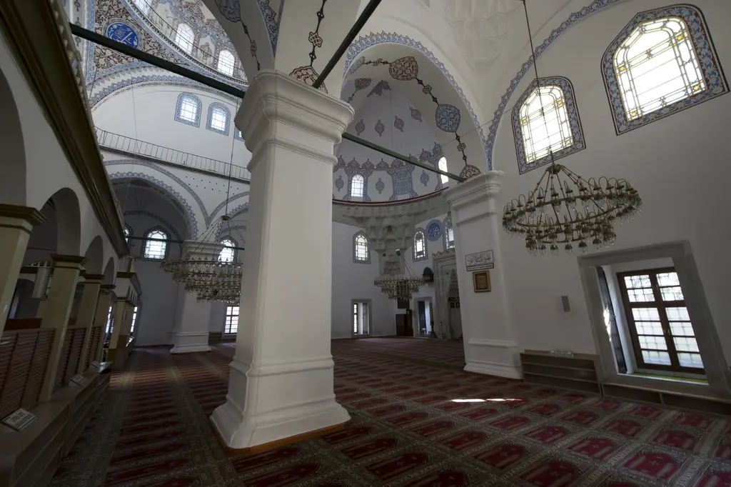 Gazi Atik Ali Paşa Camii'nde yan bölmeden harime bakış; beyaz paye, kemerler, avize, pencereler ve kırmızı desenli halı.