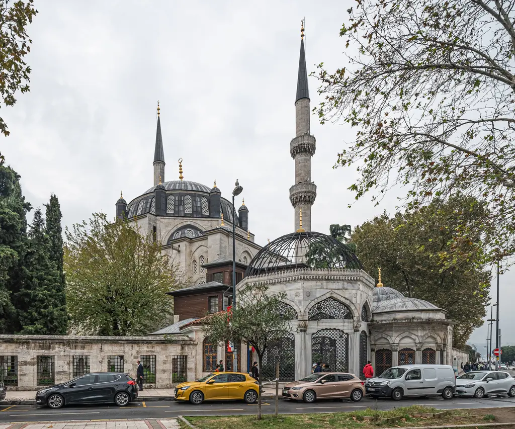 Yeni Valide Camii; ağaçların arasında kubbe ve minare, önde türbe, sebil ve caddede park etmiş arabalar.