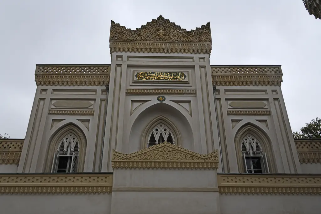 Yıldız Hamidiye Camii'nin taçkapı biçimli cephesine aşağıdan bakış; mukarnaslı taç, altın yazılı kitabe, sivri kemerli pencereler ve gri gökyüzü.