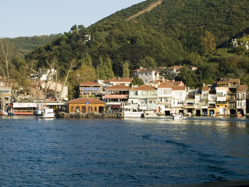 Anadolu Kavağı'nda denizden köy; beyaz ve krem evler, kıyıda lokantalar ve arkada ağaçlarla kaplı yüksek tepe.