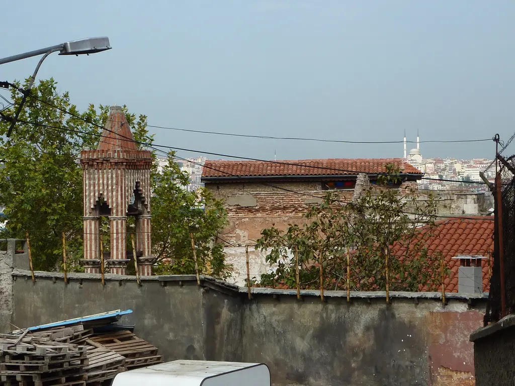 Kanlı Kilise'nin çan kulesi; tuğla sivri çatılı kule, önünde avlu duvarı, ağaçlar, kiremit çatılar ve uzakta cami minareleri.