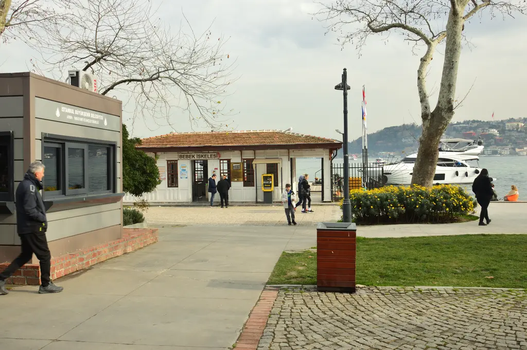 Bebek İskelesi; kiremit çatılı tek katlı iskele binası, önünde parke taşlı meydan, çimenlik ve ağaçlar.