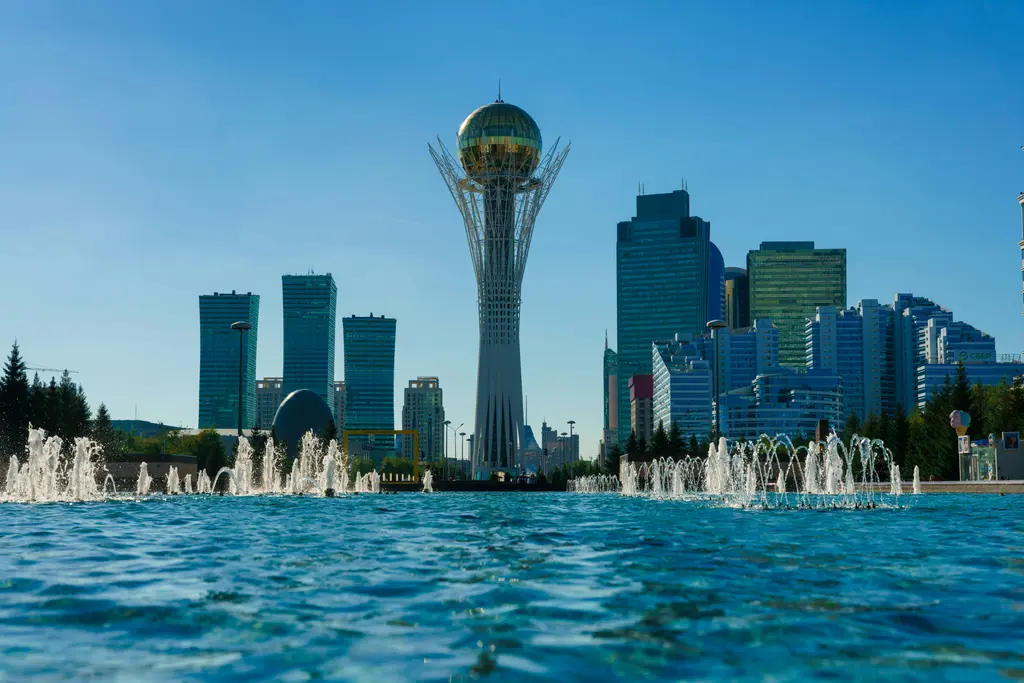 Astana'da yaz günü şehir merkezi; önde geniş havuz ile çalışan fıskiyeler, ortada Bayterek Kulesi ve iki yanında sıralanan mavi camlı gökdelenler.