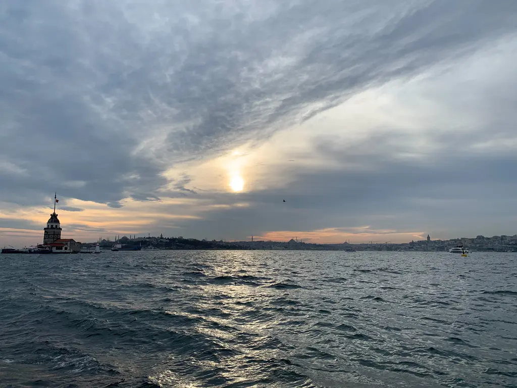 Kız Kulesi; kalın bulutların arasından görünen güneş, suya vuran ışık ve ufukta kulenin küçük silueti.