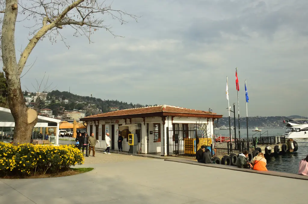 Bebek'te iskele ve kıyı; kiremit çatılı iskele binası, çıplak dallı ağaç, bayraklar ve arkada Boğaz.