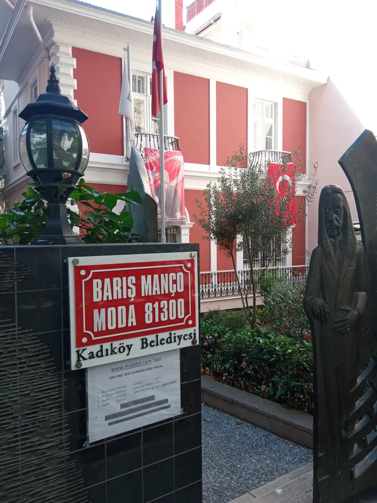 Barış Manço Evi'nin bahçe kapısı; kırmızı tabela, siyah kabartma panolar, fener ve arkada kırmızı köşk.