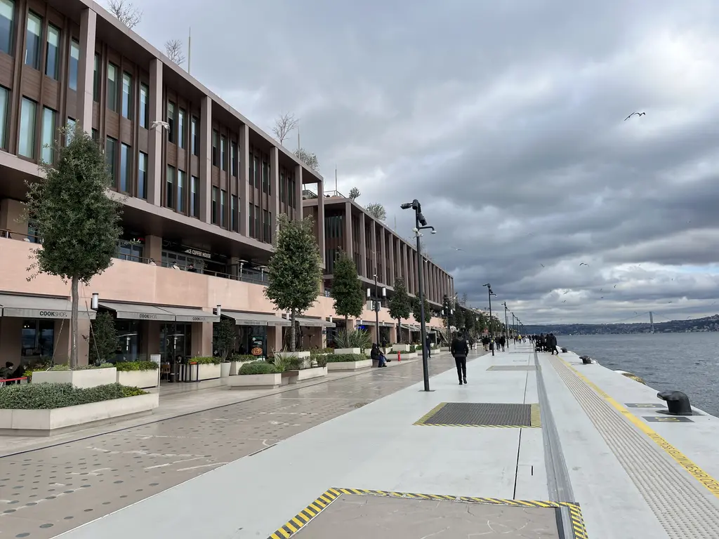 Galataport'ta kıyı yolu; solda sıra sıra ağaçlar ve tuğla rengi binalar, sağda iskele kenarı ve Boğaz, bulutlu gökyüzü.